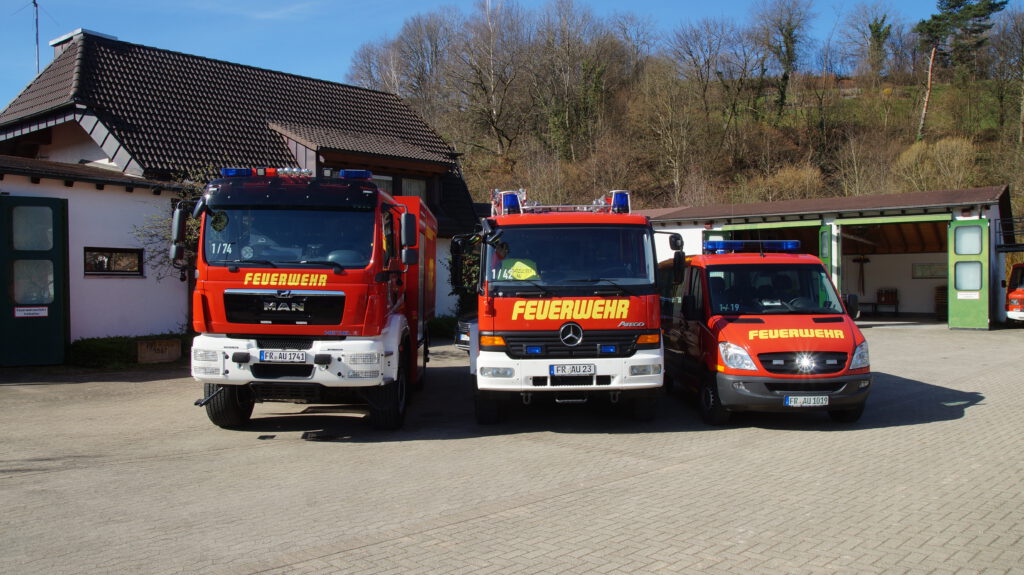 Drei Feuerwerhfarzeuge nebeneinadner, von groß nach klein. Im Hintergrund das Feuerwehrgerätehaus